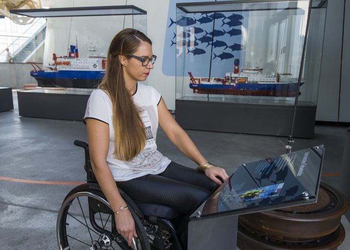 Eine Frau in einem Rollstuhl vor einer Tafel im Museum. Im Hintergrund sind Schiffsmodelle.